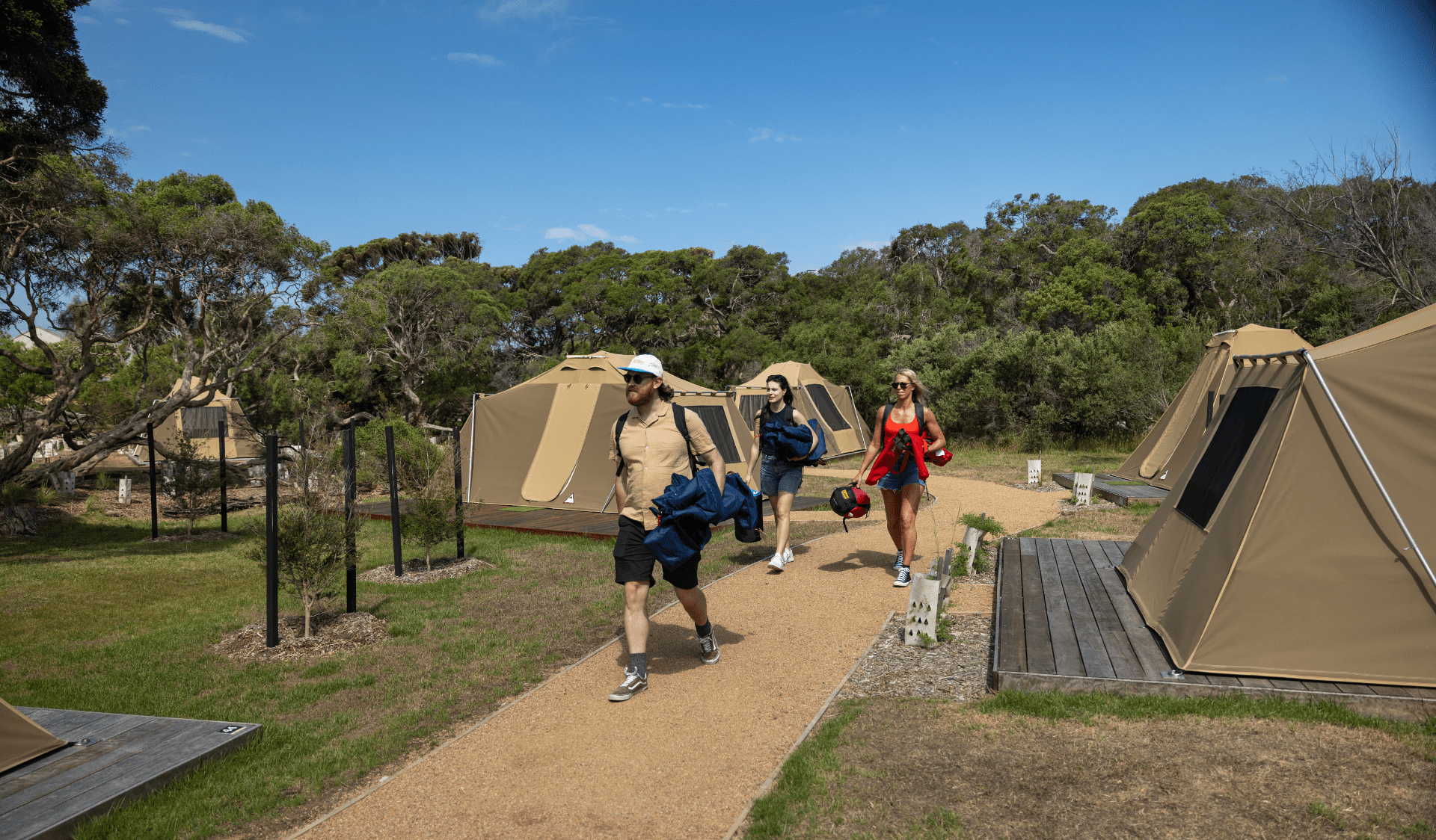 Three people walking along path near tents holding personal belongings.